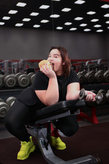 Fototapeta na wymiar Overweight girl with a sandwich and dumbbell in hand in the gym. Photo in the interior of a sports club.