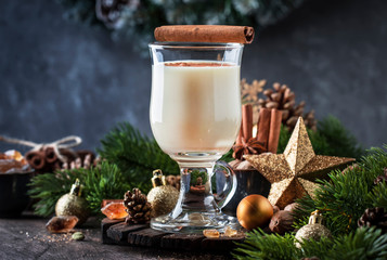 New Year or Christmas Eggnog cocktail - hot winter or autumn drink with milk, eggs and dark rum, sprinkled with cinnamon and nutmeg in a glass on wooden background, festive decoration
