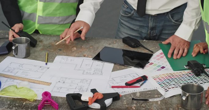 Three people surround a desk with lots of building blueprints on it and they all point at the blue prints , while one man moves a transceiver out of the way to see better , the desk is clustered with