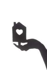 The silhouette of a female hand holding a toy house. A house with a heart-shaped window. Shadow on a white wall from sun. Creative minimal concept of home comfort.