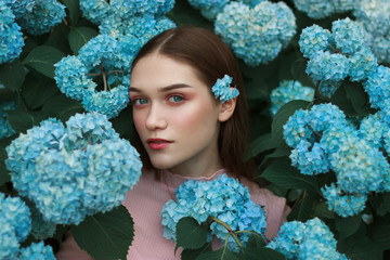 Close up face of beautiful young woman with red lips and blue eyes and with a blue flower behind her ear, standing among the flowers