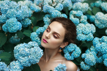 Cheerful tender young girl with healthy natural skin and red lips standing among blue flowers with naked shoulders