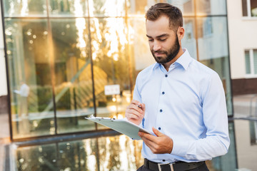 Concentrated young business man standing near business center holding clipboard writing notes.