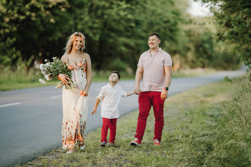 a man holds his son by a hand, and his pregnant wife stands near with flowers