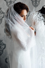 Morning of beautiful bride near the wedding dress. Bride in a white robe plays with her veil. Bride hairstyle, bride dress up at home in a robe. Nice cute girl in a boudoir robe stand near dress