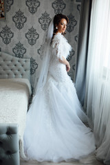 Bride morning preparation. Beautiful bride in white wedding dress and veil near  bedroom window. Adorable fiancee waiting for the groom. Young brunette bride in the morning at home.