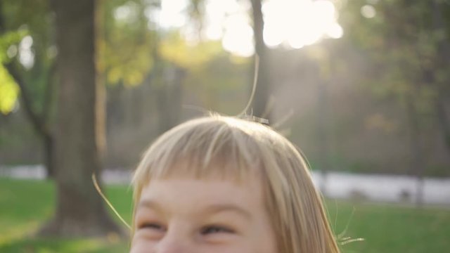 Close-up of a cute blond girl looking at the camera and laughing. Portrait of bautiful child in the park in sun rays. Leisure activity, outdoors.