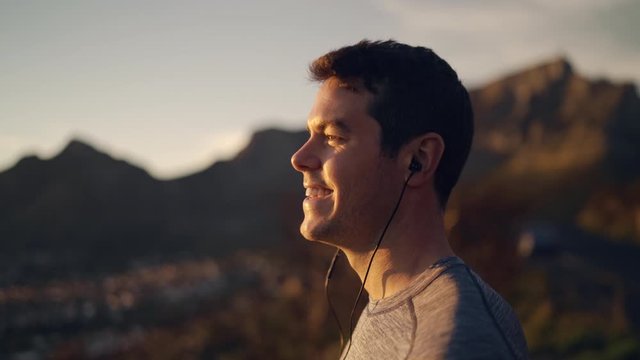 Side view of a male athlete wearing earphones enjoying the morning sunlight and cityscape view from mountain - happy man enjoying the sunrise and nature 