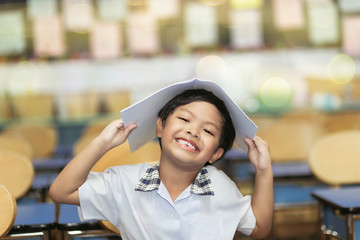An Asian boy put a book on his head with smiling sitting on a chair in a classroom.