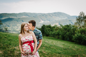 happy family. pregnant woman in a white dress with red bow, husband kiss, holds in hand round belly on the grass. The sincere tender moments. background, mountains, forests, nature, nine month