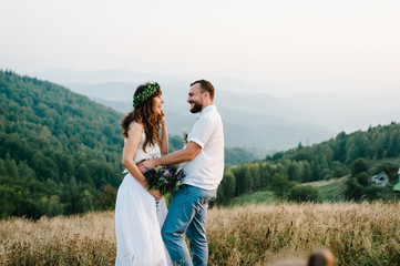 Beautiful bride in a white dress embraces the groom was in the mountains. bouquet and wreath of wild flowers. Wedding ceremony. Hipster. Rustic. Happy funny young couple. Promises talk to each other