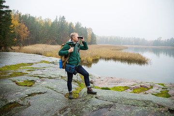 Mature man exploring Finland in the fall, looking into fog through binoculars. Hiker with big backpack standing on mossy rock. Scandinavian landscape with misty sea and autumn forest.