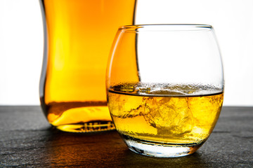 A simple and elegant whiskey (whisky) bottle, shot and glass with ice on a dark stone base with a white background with copy space