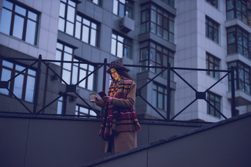 Fototapeta na wymiar Evening photo. Side view stock photo of young lady going down by stairs in the city in warm autumn clothing and using her phone texting messages. Copy space
