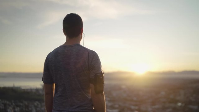Rear view of healthy sporty man looking at the city and beautiful sunrise from the mountain - cinematic inspirational sunrise 