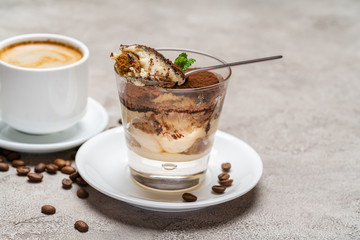 Portion of Classic tiramisu dessert in a glass and cup of coffee on concrete background