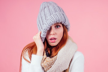 shy redhead ginger woman putting a knitted spiky hat on his head,hiding eye and open mouth in studio pink background