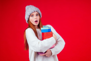 surprised redhaired ginger woman wear stylish wool hat and white sweater looking at camera and holding heavy three boxes witg gifts copyspase in studio red background