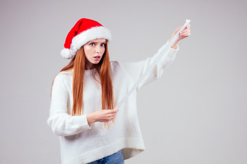 amazed and surprised redhaired ginger woman wearing red santa claus hat and knitted sweater holding long shopping receipt list in studio white background. big money xmas spending