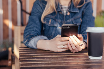 Fototapeta na wymiar Unidentified young girl in casual clothes chatting in social networks using a smartphone while sitting at the wooden table of an open cafe in the warm summer time