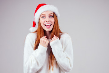 beautiful redhaired ginger woman wearing white knitted sweater and santa claus hat finger cross making wishes in studio background