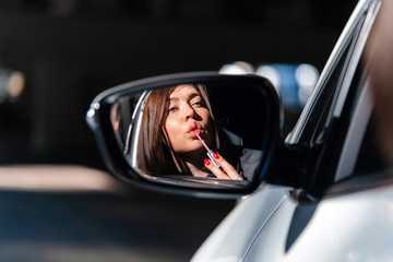 Confident and beautiful young business woman painting her lips in new car and looking into Side view mirror.