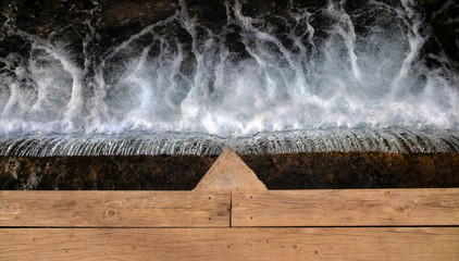 Puente pasarela de madera sobre río y pequeña cascada de agua provocando espuma