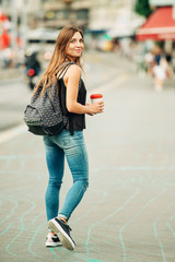 Outdoor portrait of beautiful young woman walking down the road in the city, wearing backpack,...