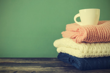 Bunch of knitted pastel color sweaters with different knitting patterns perfectly folded in stack on brown wooden table, green textured background. Fall winter season knitwear. Close up, copy space.