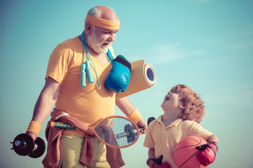 Senior man and cute little boy exercising on blue sky background - isolated. Age is no excuse to...