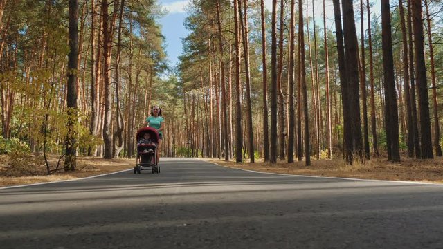 Active athletic woman running with a baby stroller on an asphalt road through a pine forest. A girl runs past the camera. Active family with baby jogger. Slow-motion 4k shot