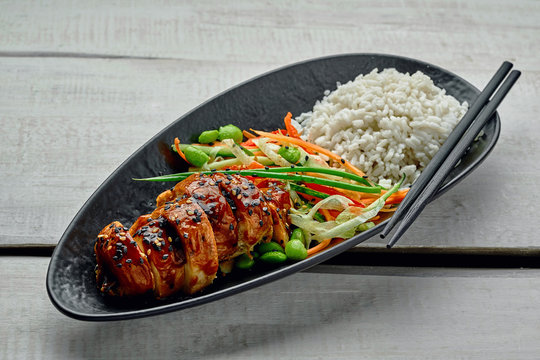 Asian- style chicken with savory and sweet teriyaki sauce, rice, sesame, salad, edamame and sweet chili sauce on a white wooden table.