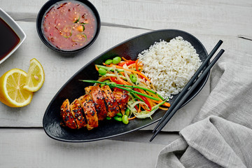 Traditional Japanese teriyaki chicken with cucumber, ginger, carrot, pepper and cabbage salad, rice, sesame and edamame, lemon, chili and soy sauces on a rustic table. Top view, directly above.