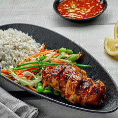 Chicken with savory and sweet teriyaki sauce, cucumber, cabbage, ginger and carrot salad, rice, sesame and edamame in a black pottery dish.