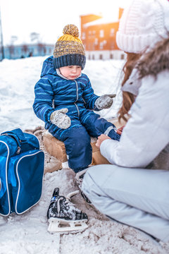little boy of 3.5 years old, winter on city skating rink, sits on bench, woman s mother changes her child s clothes, puts on shoes, rest on weekend, snow drifts, warm clothes, hats, jackets.