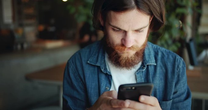 Young red-bearded male hipster typing message on smartphone in cafe, using modern technologies