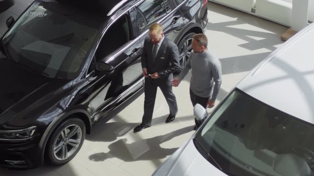 High angle shot of professional salesman walking with male customer in auto showroom and discussing cars