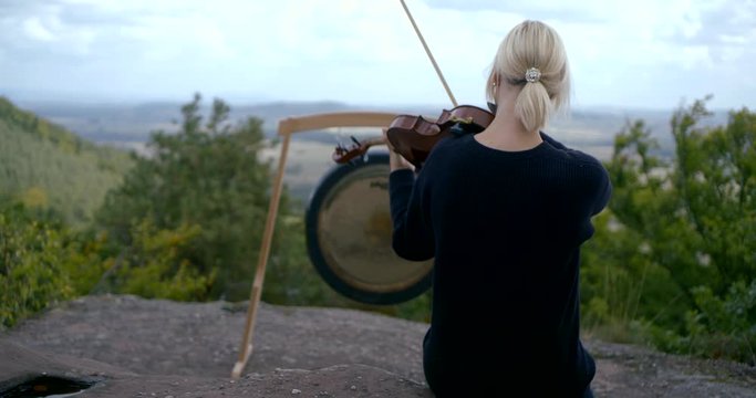 Blonde girl playing violin sitting on a mountain in the background of space and Gong. Eastern medicine treatment of musical vibrations