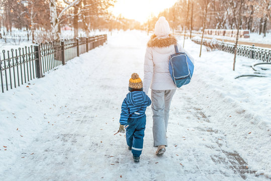 Woman Mother Walks Down Street Winter, With Small Child, Boy Or Girl 3-5 Years Old, View From Rear, Background Road Snowdrifts, Returns Kindergarten School Workout. Holiday Weekend Warm Clothes.