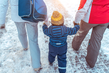 Woman mother walks winter along street with her friend, with small child boy or girl 3-5 years old, view from back, background road snowdrifts, returns from kindergarten school, warm clothes hats.