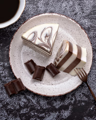 Cup of coffee and delicious jelly dessert with white & brown layers, milky glaze and pieces of dark chocolate on a spotty plate. Dessert spoon. Delicacy. Nice black background. Top view.