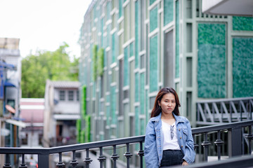 Portrait of Hipster Asian woman in white t-shirt, jean jacket and black trousers with beautiful modern building in background