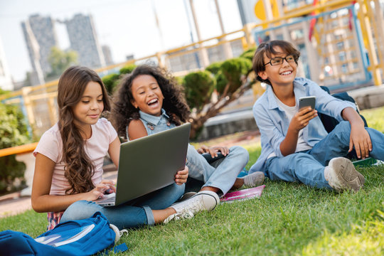 Group of multiethnic elementary school children sitting on playground grass looking at laptop computer