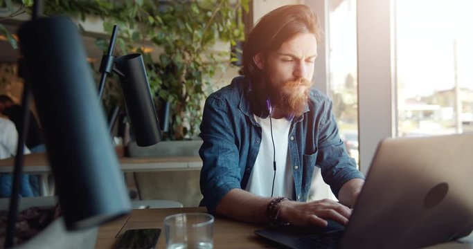 Attentive man in casual clothing working on laptop in cafe, sitting near window, journalist