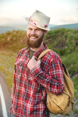 Naklejka na ściany i meble Portrait of a bearded happy smiling traveler hipster with a backpack in a plaid shirt and a hat next to an unknown car stands on the road at sunset in the mountains. Happy and confident travel concept