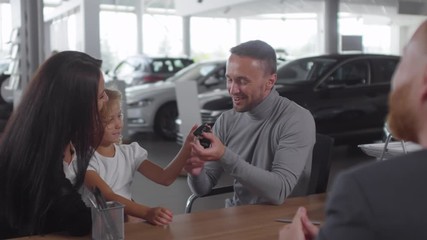 Cheerful man taking car key and shaking hands with salesman while sitting together with beautiful wife and little daughter and buying new auto in dealership center