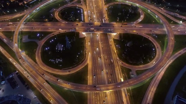 Cinematic flight over clover leaf road  night complex intersection in modern metropolis. Many transport cars ride freeway. Logistics driving taxi delivery. Aerial top down forward