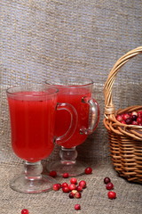 Basket filled with cranberries. It stands on a surface covered with linen cloth. Next to the glasses are cranberry juice. Several berries are scattered on the surface.