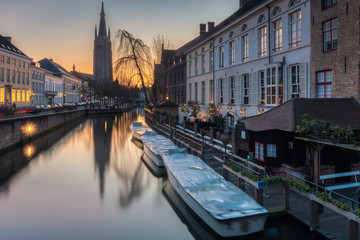 Fototapeta premium Church of our lady with traditional building facade reflects on still canal with tourist boats foreground during twilight at Bruges, Belgium
