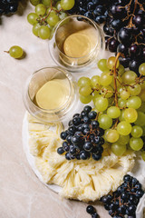 Swiss cheese tete de moine on ceramic board, black and green grapes and two glasses of white wine, served on pink marble background. Flat lay, space